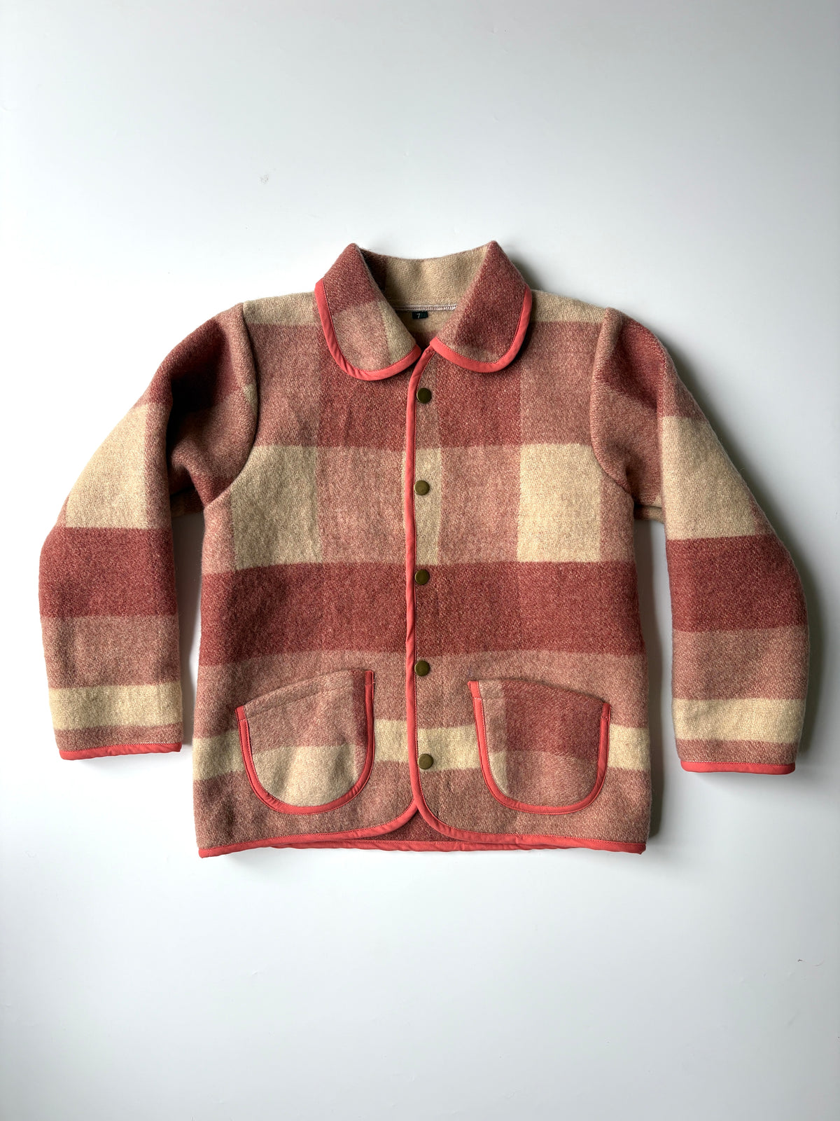 A red and cream plaid blanket children's jacket, featuring a Peter Pan collar, button closure, and two front patch pockets with red piping, laid flat against a light gray background.