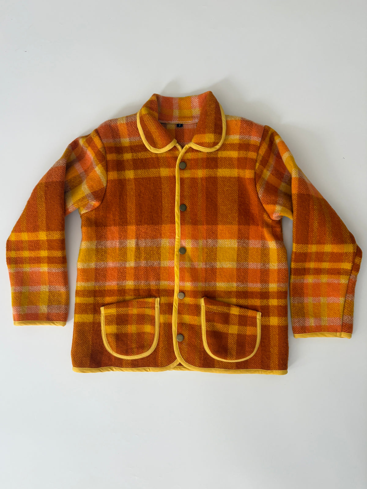 Orange and yellow plaid blanket children's jacket, featuring a Peter Pan collar, yellow piping, and two front patch pockets, laid flat on a white background.