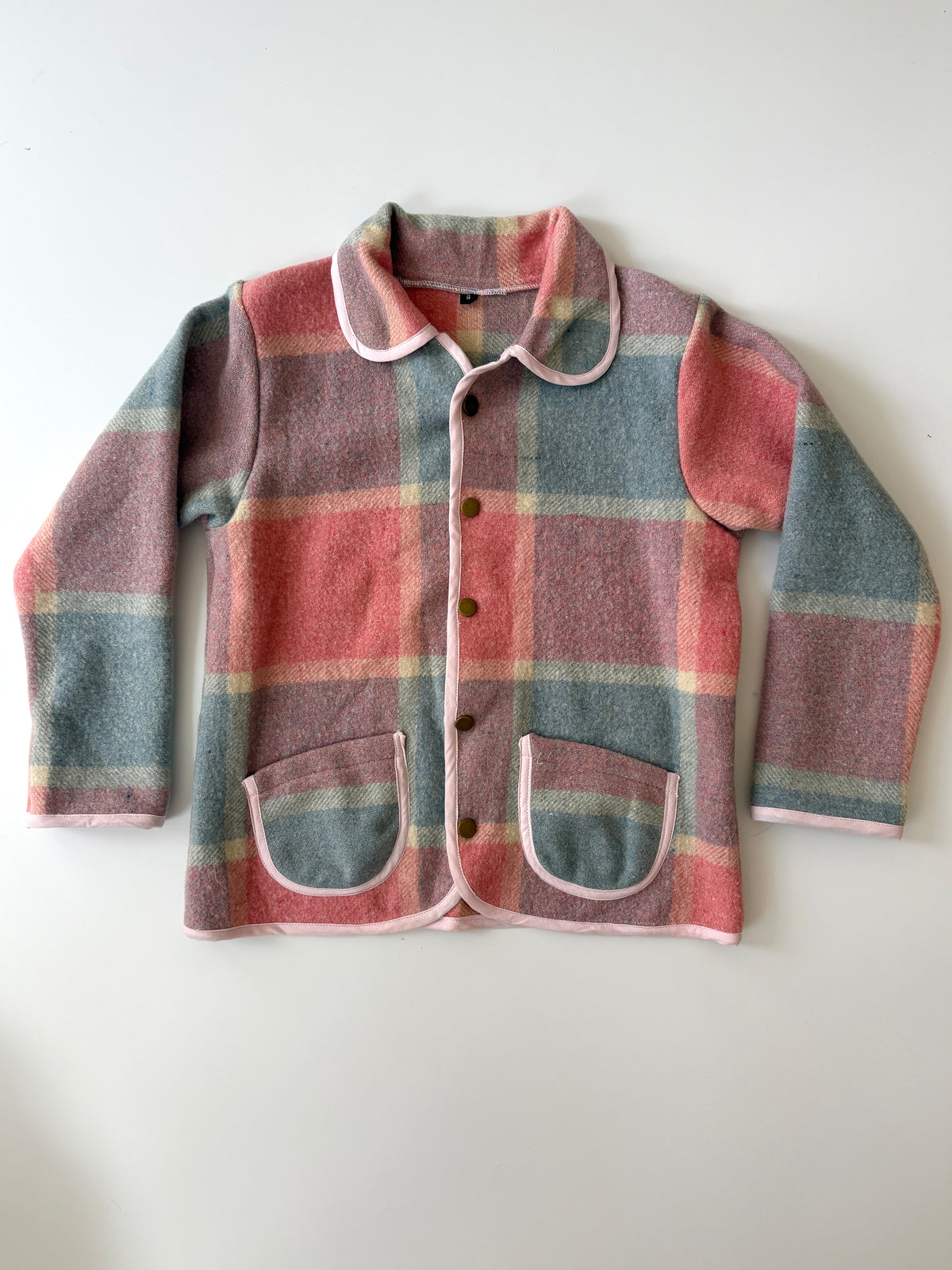 A children's long-sleeve jacket with a pink, blue, and beige plaid pattern, featuring a Peter Pan collar, button front, and two front patch pockets, laid flat against a light background.