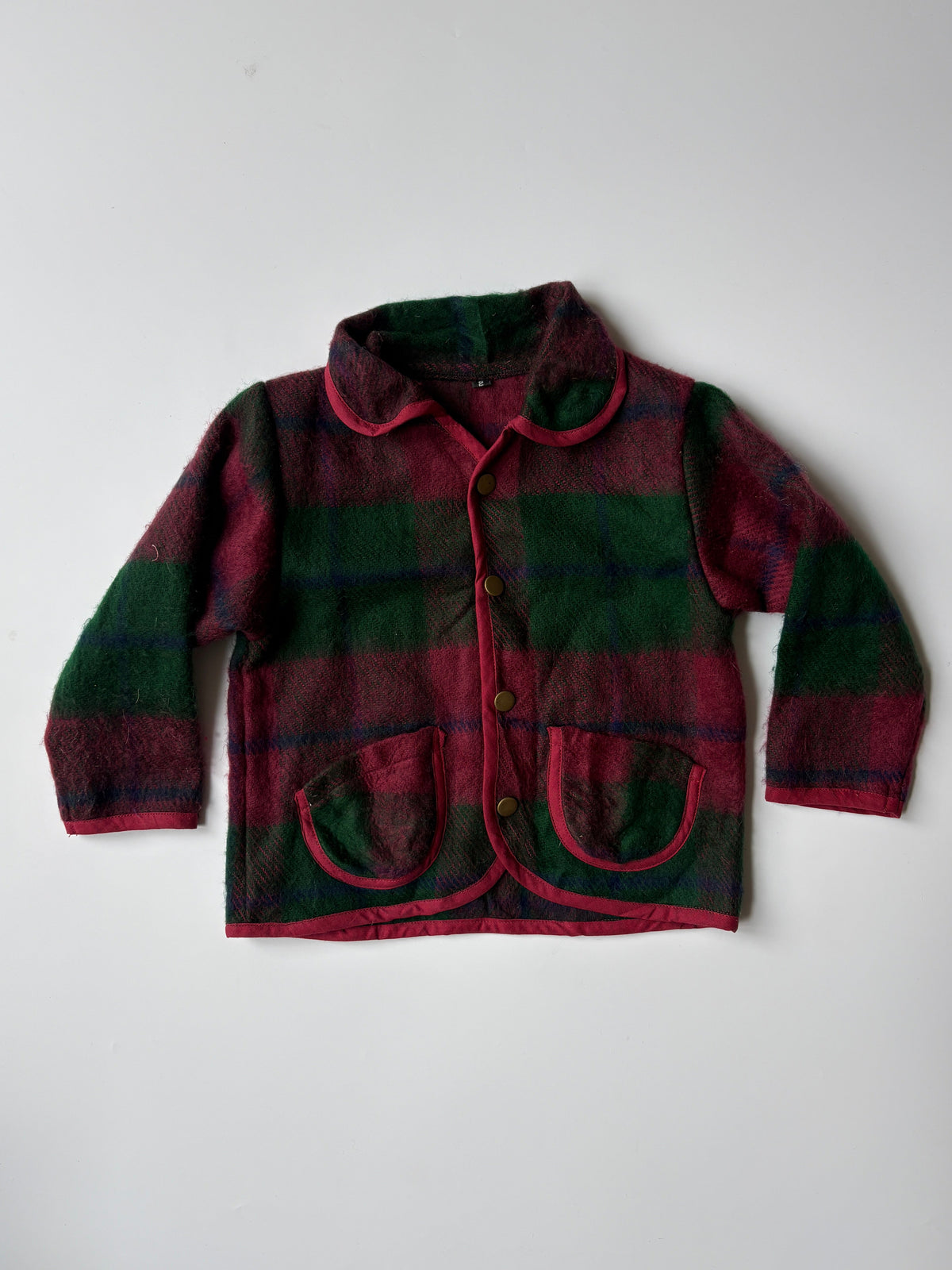Red and green plaid blanket children's jacket, featuring a collar, button front, and two front pockets, laid flat against a light gray background.