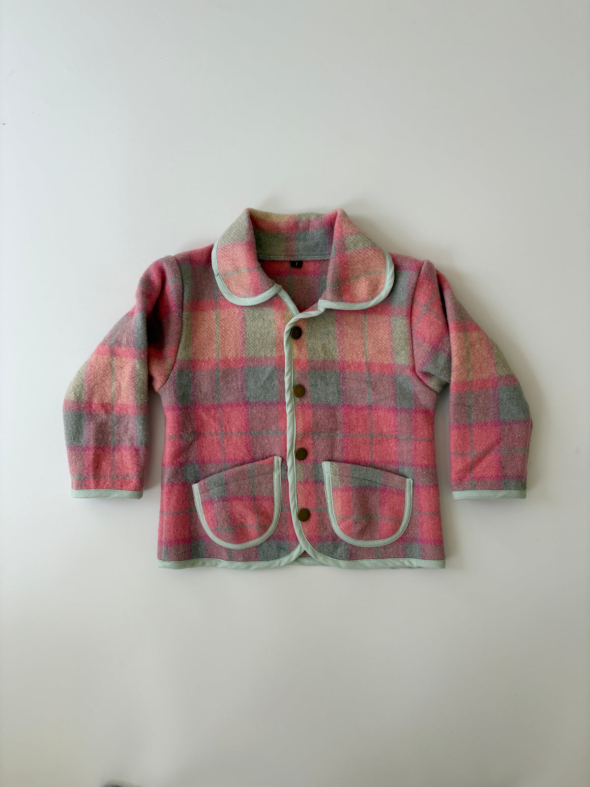 An upcycled children's plaid jacket in pink, red, and gray wool, featuring a Peter Pan collar and pocket trim, laid flat against a neutral off-white background.