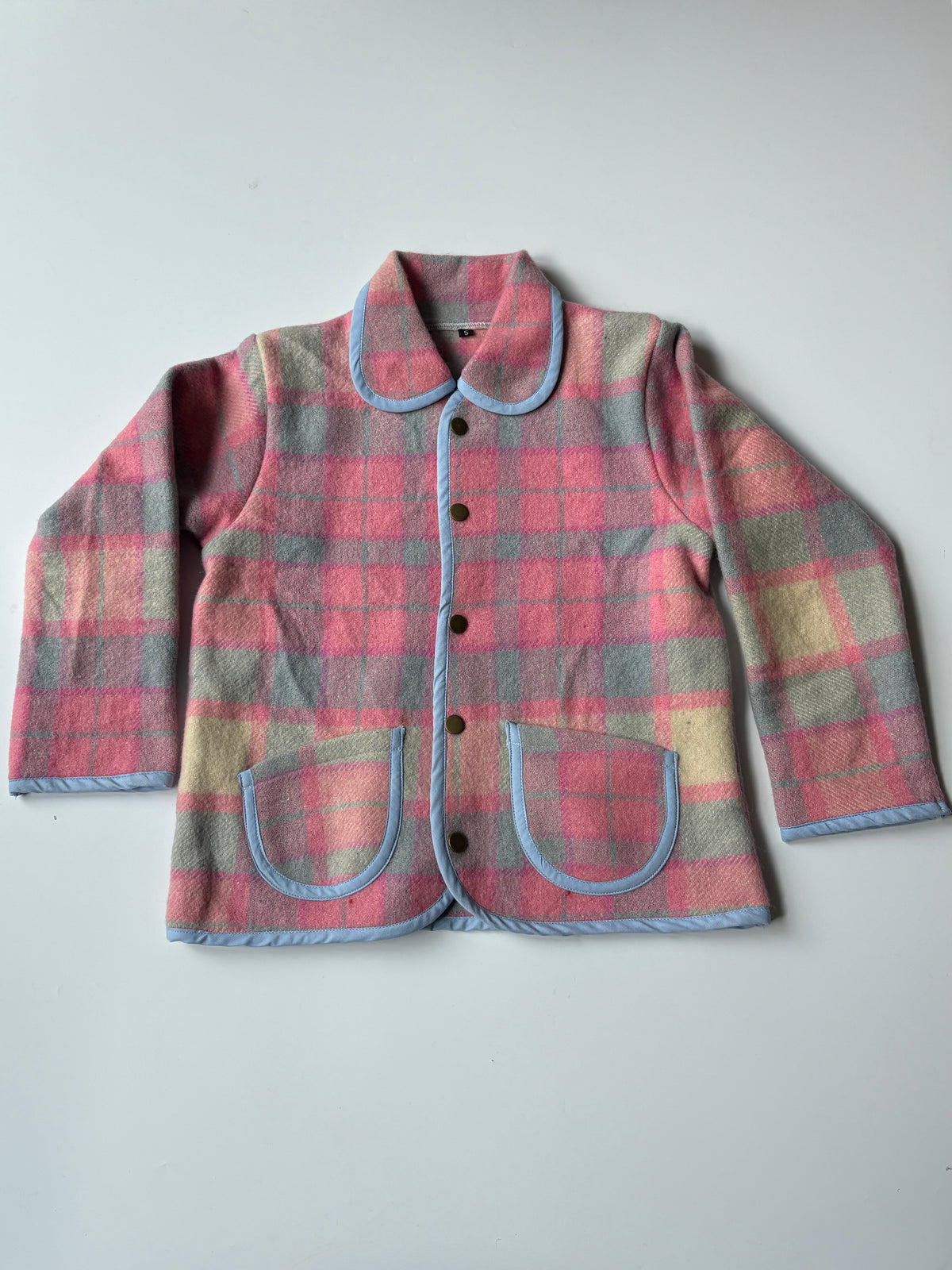 Children's button-front jacket in a pink, blue, and beige plaid pattern, featuring light blue trim on the collar, pockets, and edges, laid flat on a light gray background.