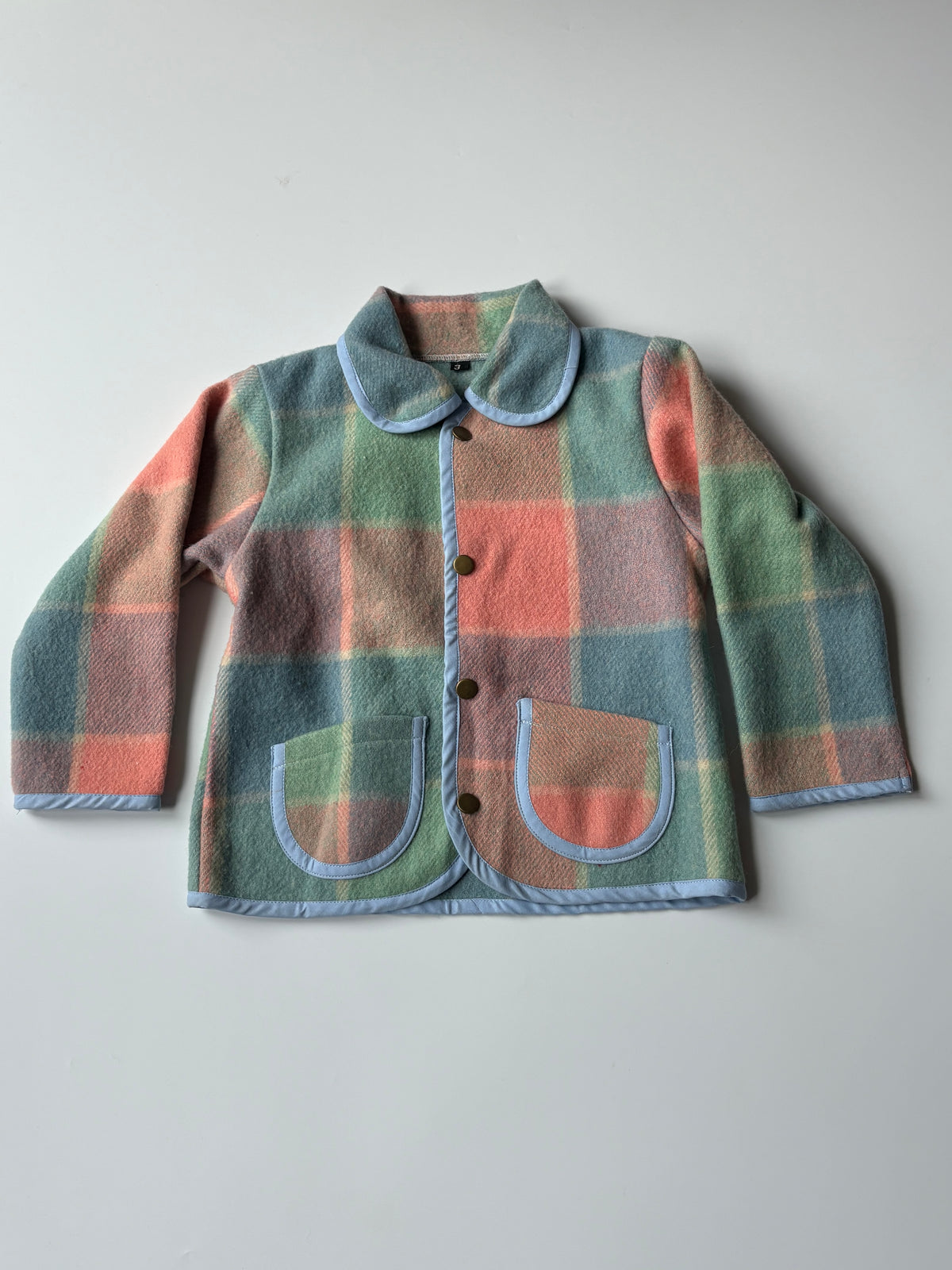 Children's pink blue green blanket jacket. A children's long-sleeve jacket in a muted plaid pattern of sage green, dusty pink, and light blue, featuring a light blue collar and trim, laid flat on a neutral gray background.