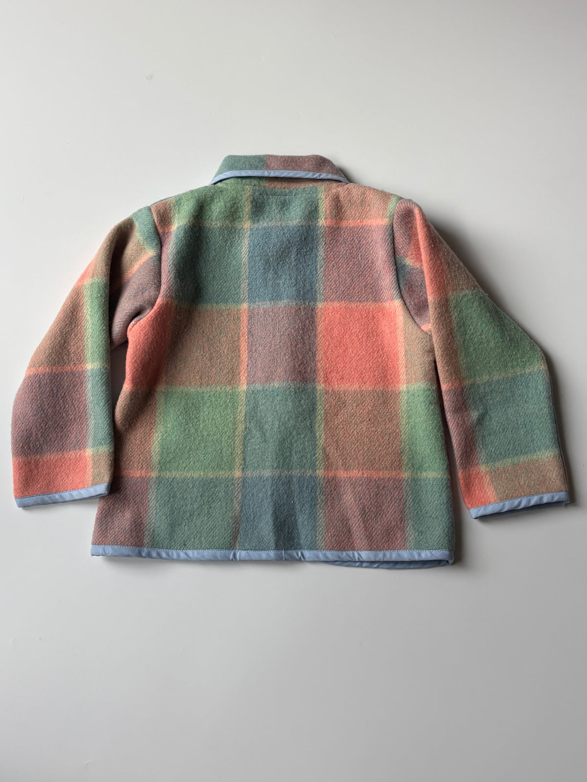 Children's pink blue green blanket jacket. A children's long-sleeve jacket in a muted plaid pattern of sage green, dusty pink, and light blue, featuring a light blue collar and trim, laid flat on a neutral gray background.
