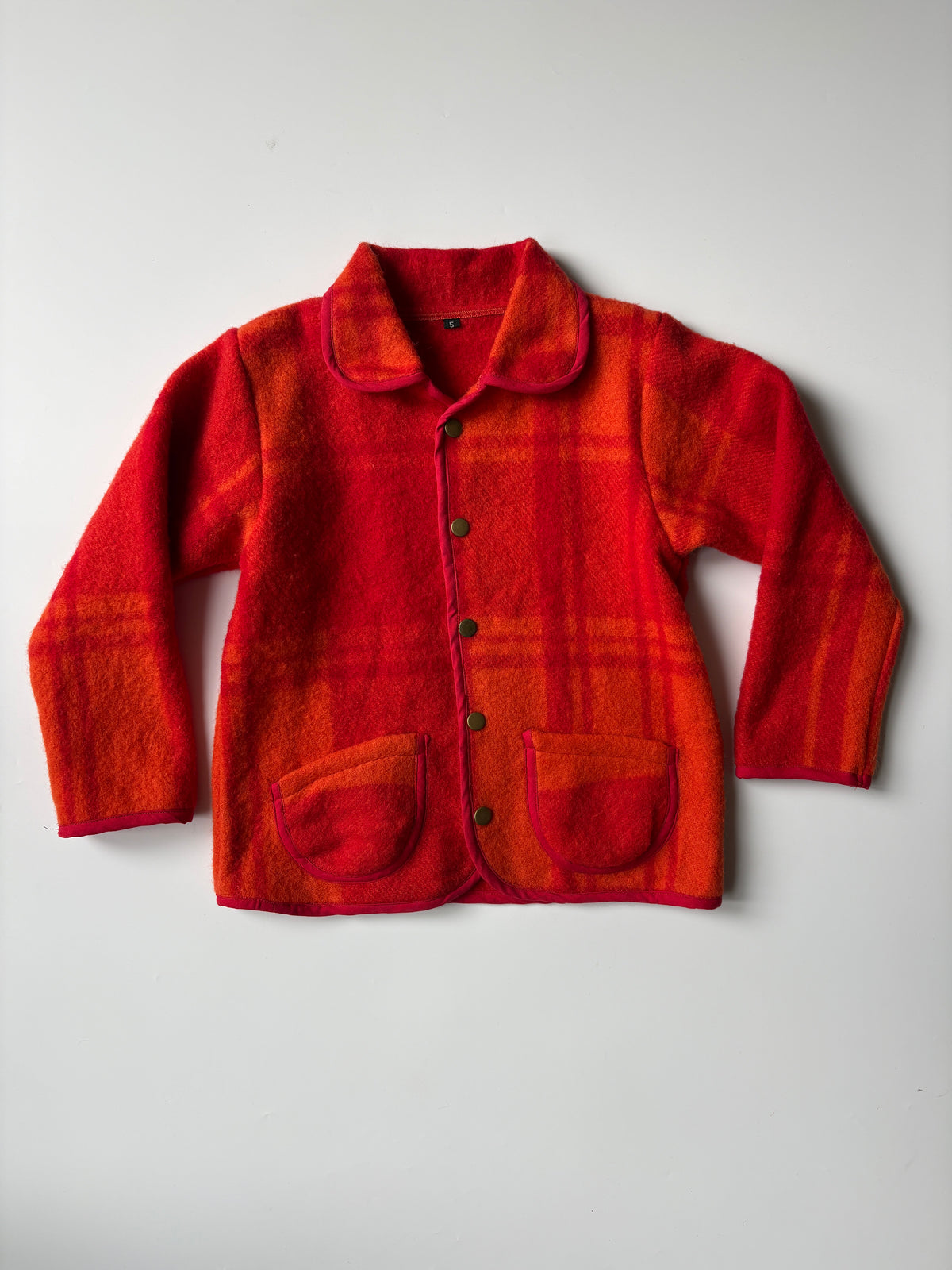 Orange & red plaid blanket children's jacket with a button front and two front patch pockets, laid flat on a light gray background.