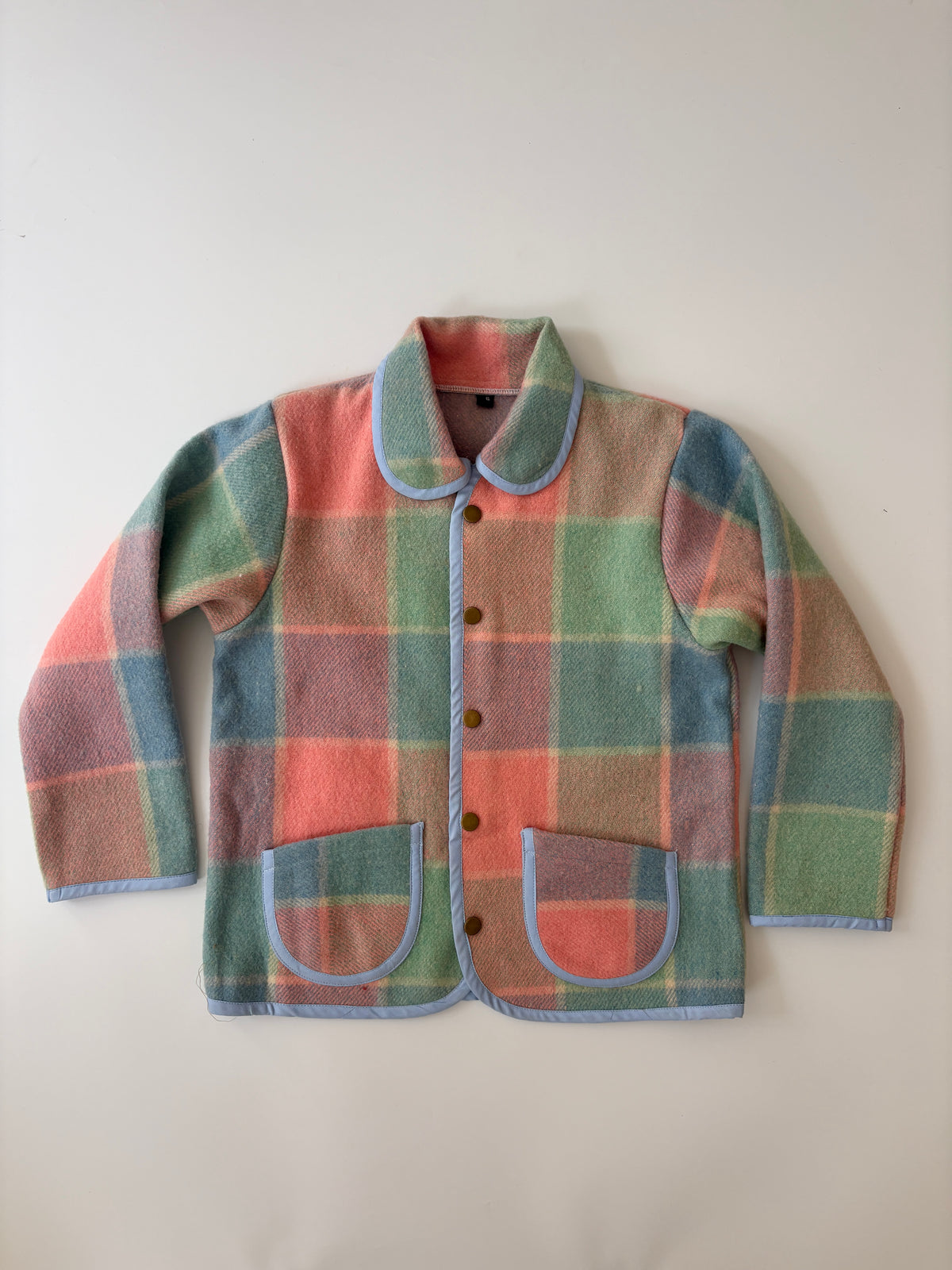 Pink green and blue blanket plaid children's jacket. A vintage-style plaid jacket with a pastel color scheme of green, pink, and blue, featuring a Peter Pan collar and front button closure, laid flat against a neutral background.