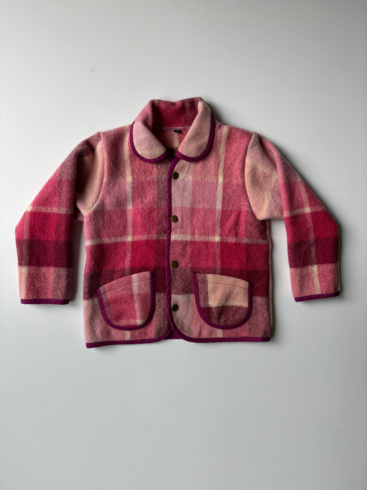 A pink and red plaid blanket children's jacket with a Peter Pan collar and front buttons, laid flat on a light gray background.