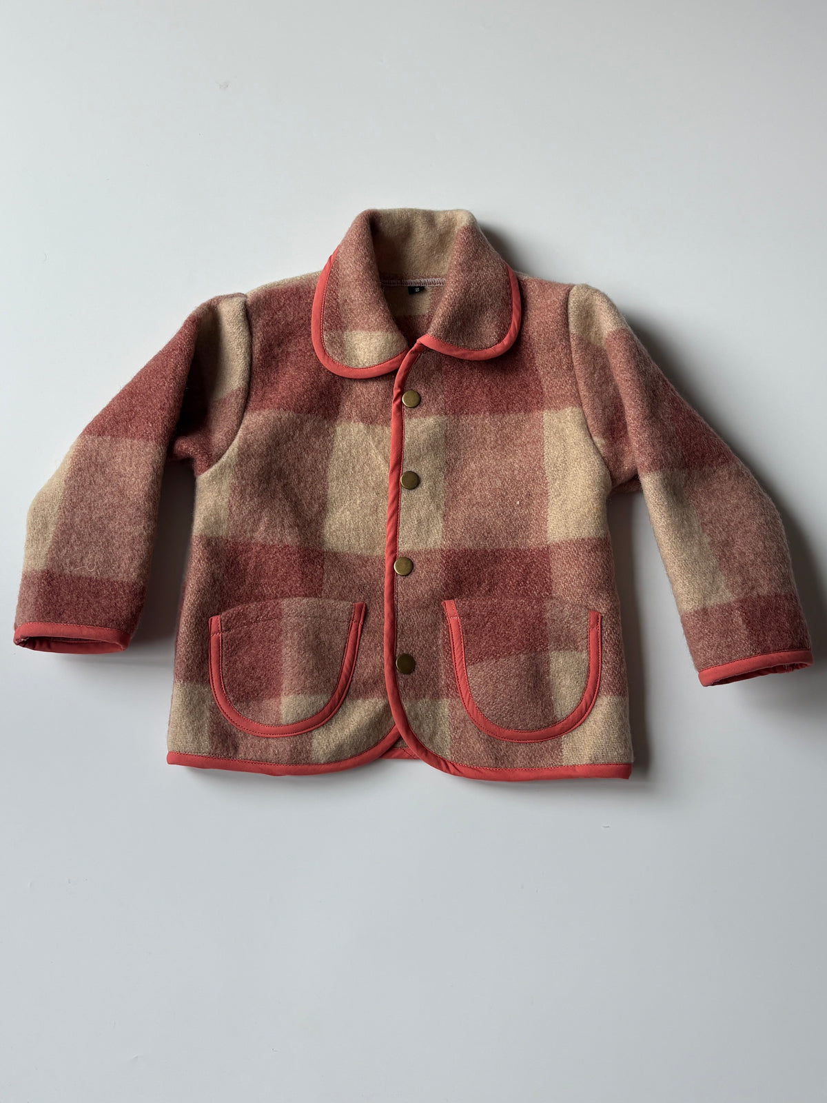 Red and cream plaid blanket children's jacket 1. A children's plaid jacket in muted red and cream with contrasting red trim, shown flat against a light gray background.