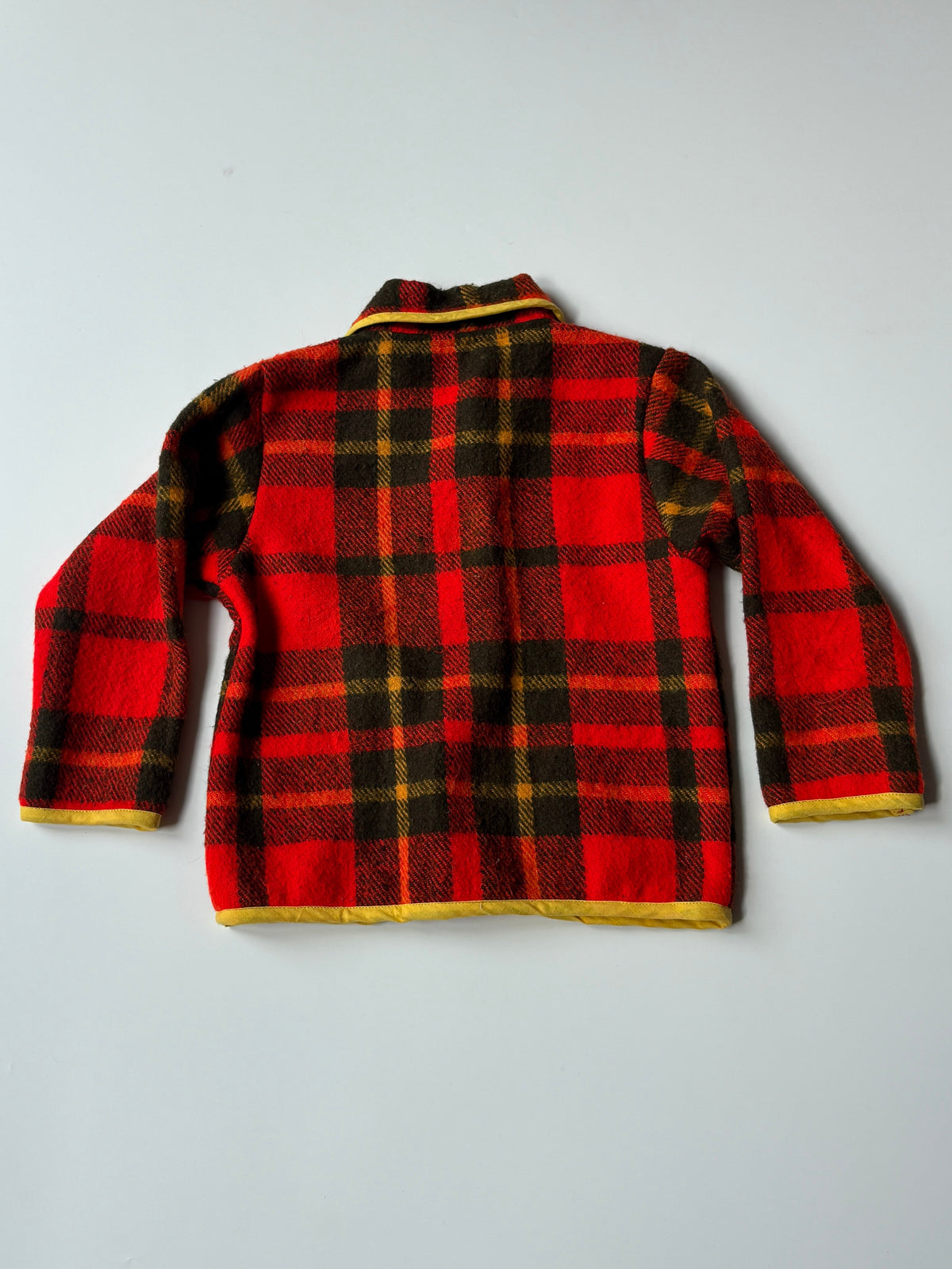 Red yellow and black plaid blanket children's jacket with yellow trim, featuring a collar, snap-button front, and two front patch pockets, laid flat on a light gray background.