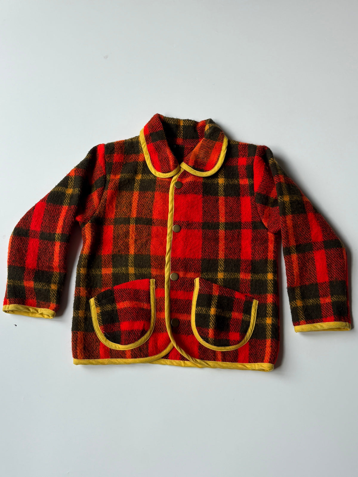 Red yellow and black plaid blanket children's jacket with yellow trim, featuring a collar, snap-button front, and two front patch pockets, laid flat on a light gray background.