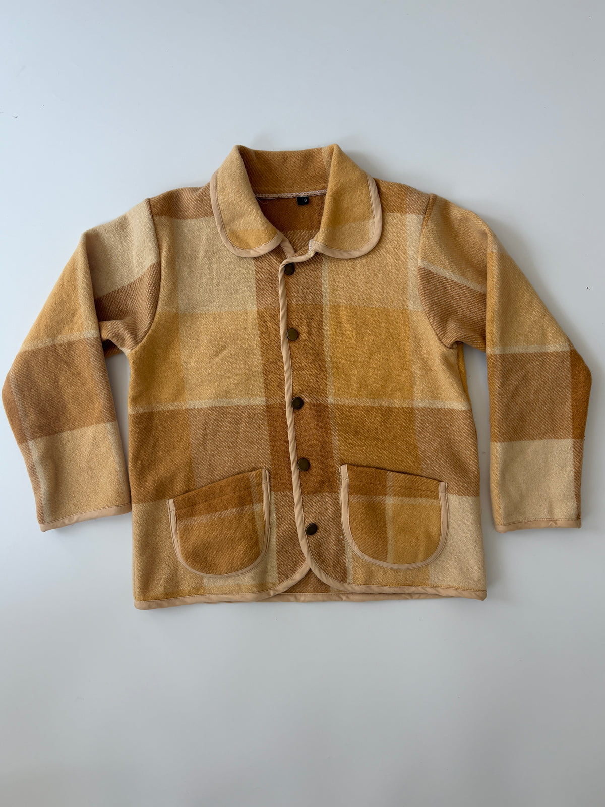 Tan and Brown plaid children's blanket jacket. A children's long-sleeve button-front jacket in a warm tan and brown plaid fabric, featuring a collar, dark buttons, and two front patch pockets, laid flat on a light gray background.