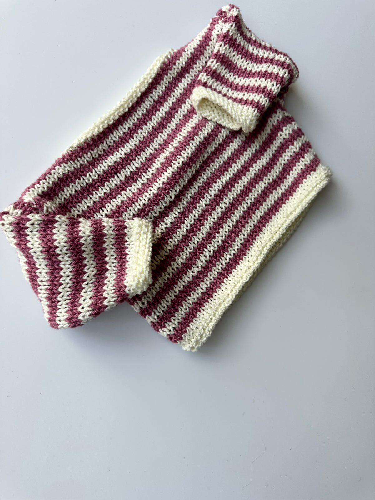 A hand-knit striped jumper in pink and white, laid flat on a light gray background.