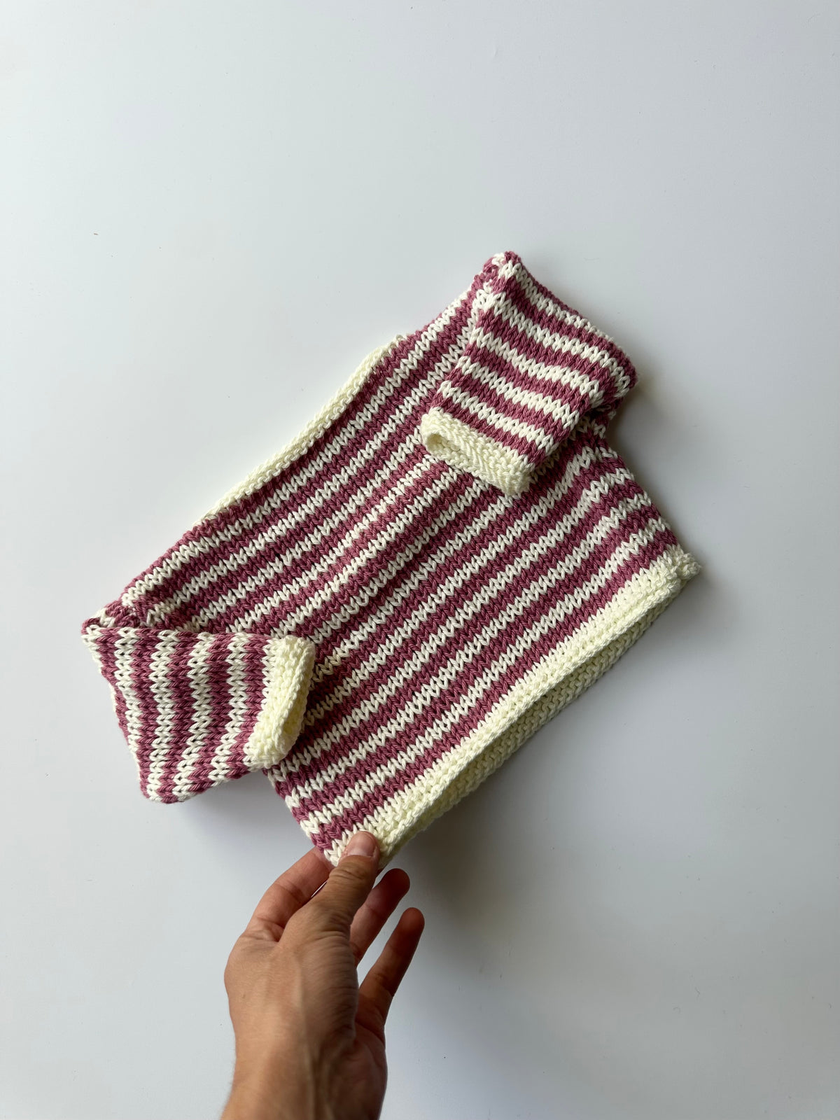 A hand-knit striped jumper in pink and white, laid flat on a light gray background.