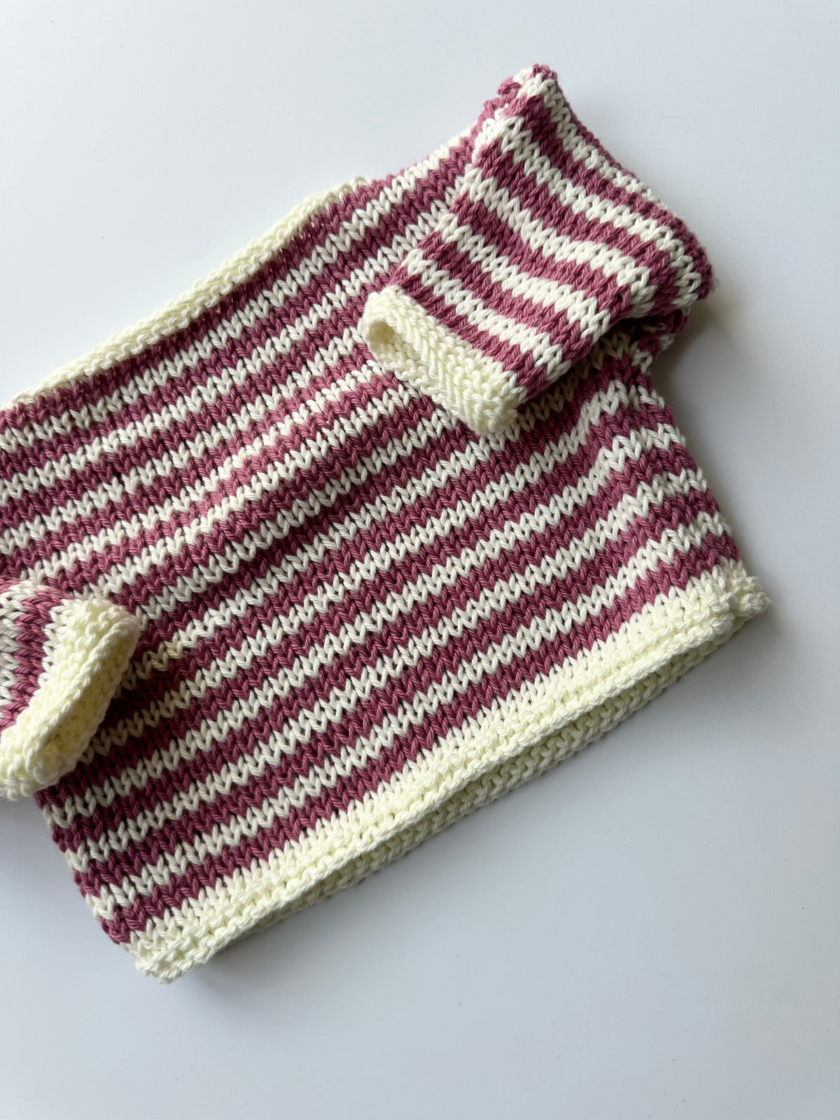 A hand-knit striped jumper in pink and white, laid flat on a light gray background.