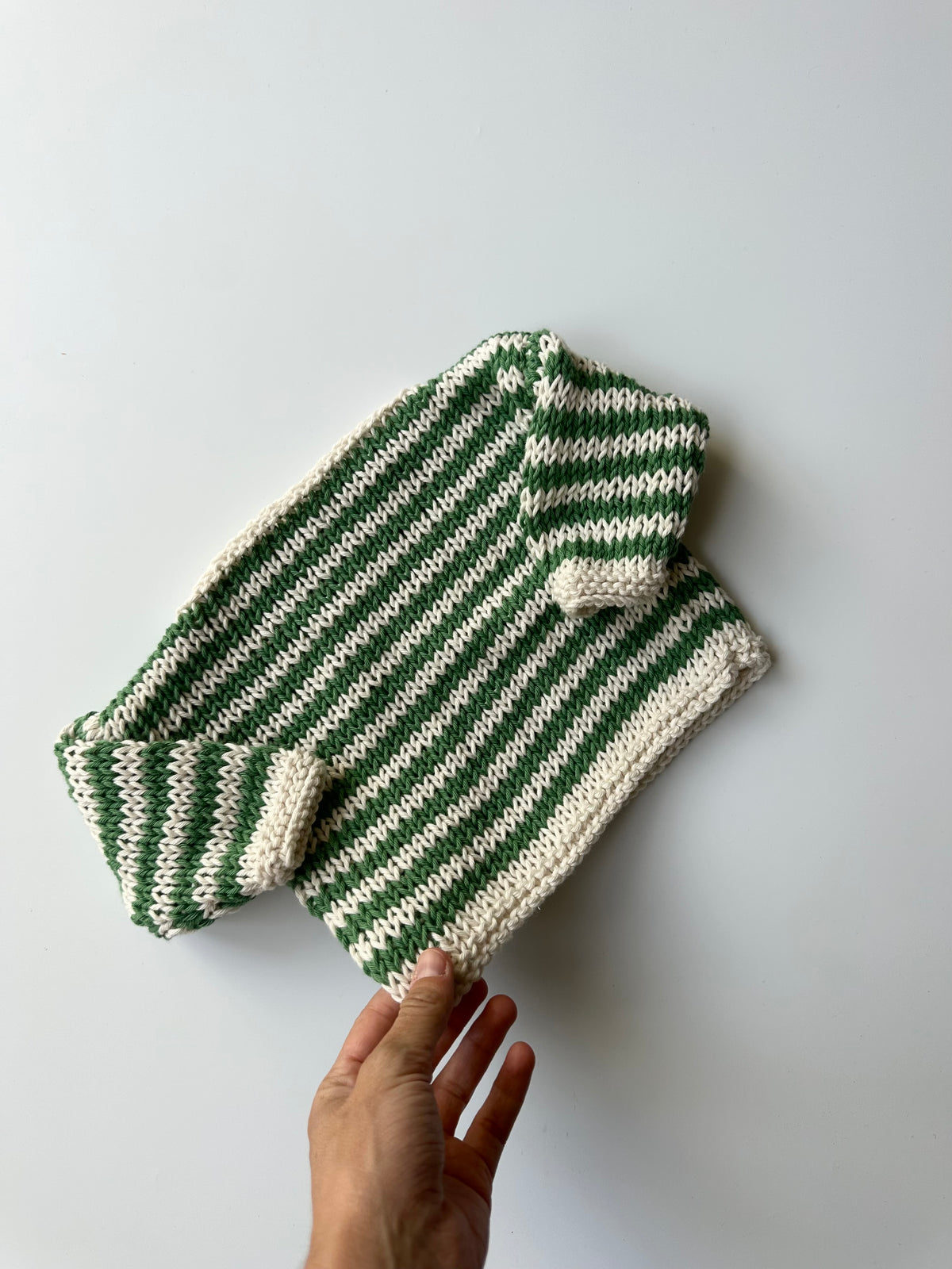A hand-knit striped sweater in green and cream, lying flat on a light gray background.